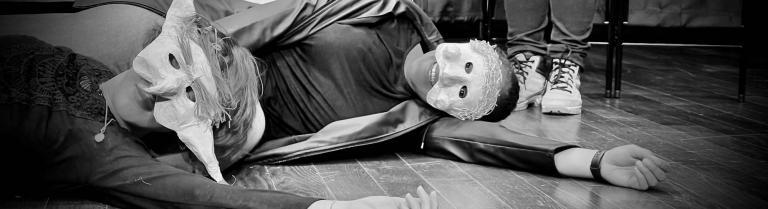Zwei Theaterschauspielende mit Masken liegen auf dem Boden