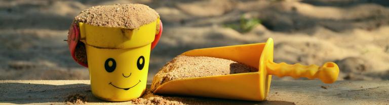 Gelbes Sandeimerchen mit lächelndem Gesicht, gefüllt mit Sand. Rechts neben  dem Eimerchen liegt eine kleine gelbe Sandschaufel