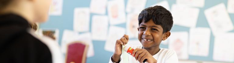 Ein Kind, welches einen Leimstift in der Hand hat, lacht mit der Lehrerin