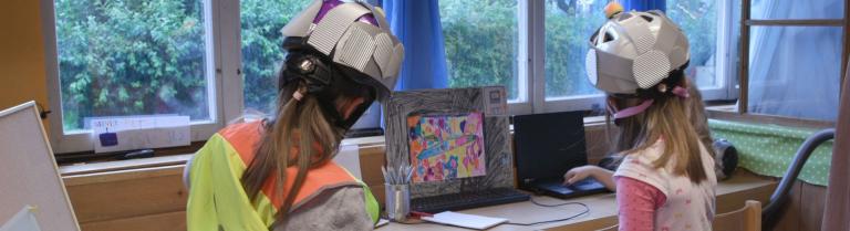 Zwei Mädchen mit Warnweste und Helm auf dem Kopf spielen Roboter im Kindergarten