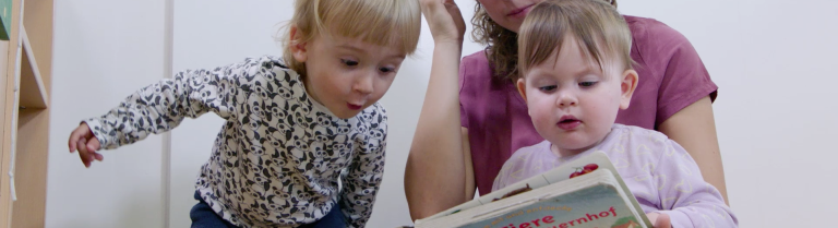 Eine Betreuerin betrachtet ein Bilderbuch mit zwei Kleinkindern