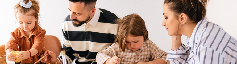 Zwei erwachsene Personen sitzen mit zwei Kindern am Tisch und malen