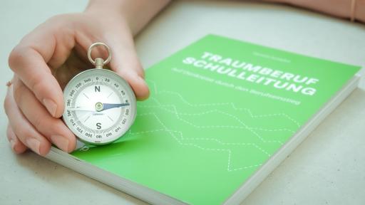 in der Hand ein Kompass auf einem grünen Buch zum Thema Schulleitung