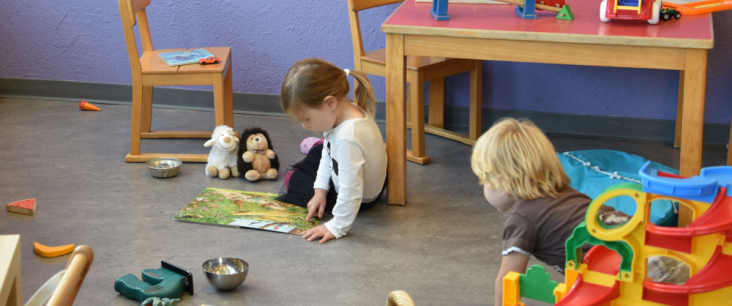 Mädchen betrachtet am Boden sitzend Bilderbuch mit Plüschtieren