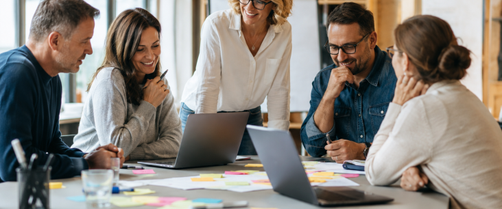 Fünf Erwachsene arbeiten lachend gemeinsam an einem Tisch mit Laptops und Post-its als Symbol für kollaborative Schulentwicklung
