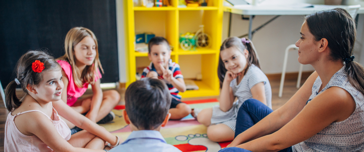 Erwachsene Person sitzt mit Kindern in einem Kreis am Boden