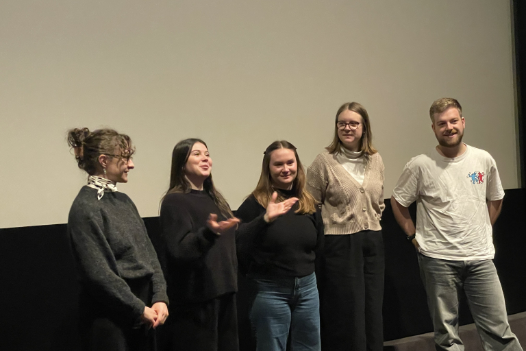 Vier Personen stehen auf der Kinobühne vor der Leinwand