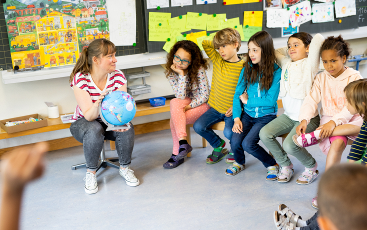 Lehrperson mit Globus sitzt im Kreis mit Kindern