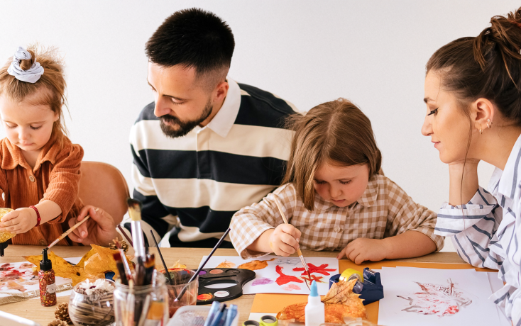 Zwei erwachsene Personen sitzen mit zwei Kindern am Tisch und malen