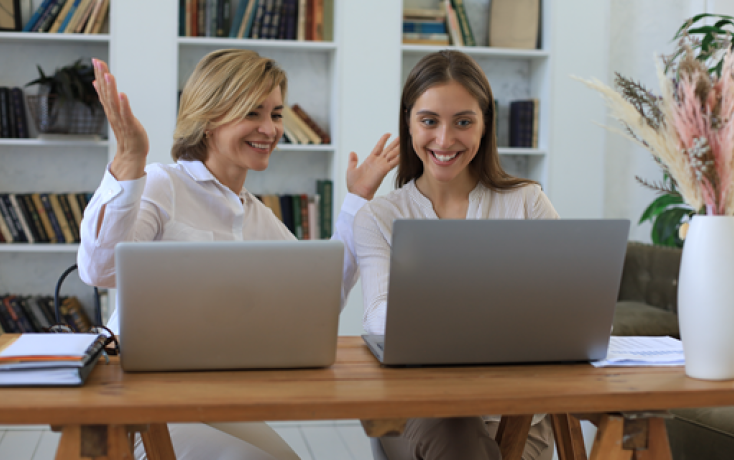 Zwei Frauen sitzen lachend vor Laptops und beraten sich gegenseitig zu ICT-Themen