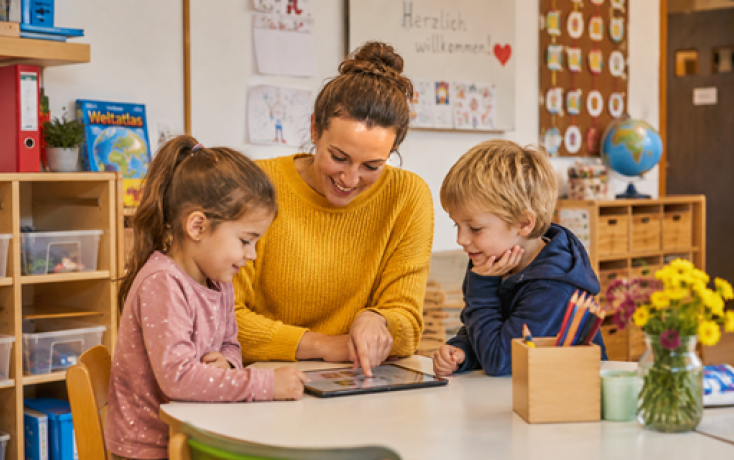 Lehrperson zeigt zwei Kindergartenkindern etwas auf einem Tablet in einem farbenfrohen Klassenzimmer