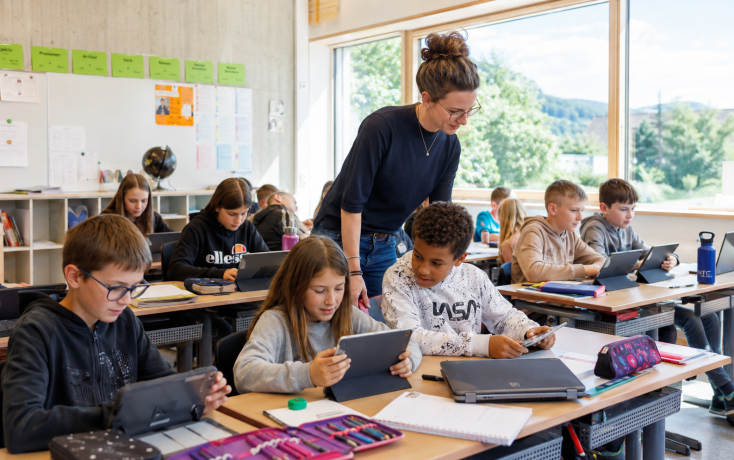 Lehrperson unterstützt Schülerinnen und Schüler beim Arbeiten mit Tablets in einem modernen Klassenzimmer