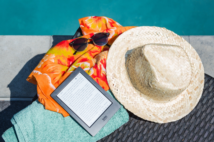 Ein Strandtuch, ein E-Reader, eine Sonnenbrille und ein Strohhut liegen neben einem Pool.