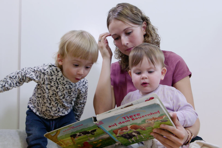Eine Betreuerin betrachtet ein Bilderbuch mit zwei Kleinkindern