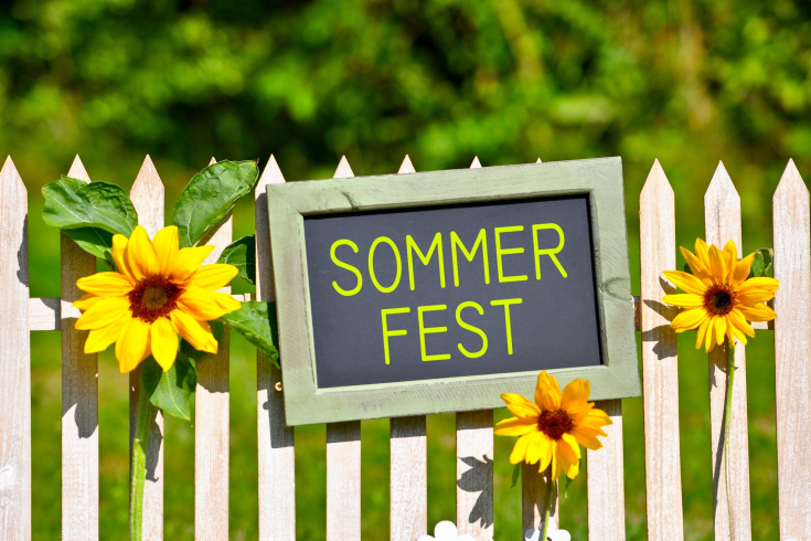 Ein Schild hängt an einem Gartenzaun. Darauf steht: "Sommerfest"